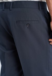 Mann iført marineblå dressbukser med knappet baklomme og beltestropper, med hånden hvilende nær lommen.