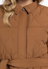 Vêtement extérieur en tissu matelassé marron avec col, fermeture par boutons-pression et ceinture à la taille. Présente des motifs de couture incurvés subtils.