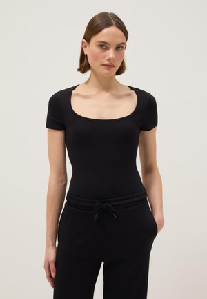 Jonge vrouw met kort bruin haar, gekleed in een aansluitend zwart T-shirt met korte mouwen en een ronde hals, en zwarte broek met trekkoord, staand tegen een effen achtergrond.