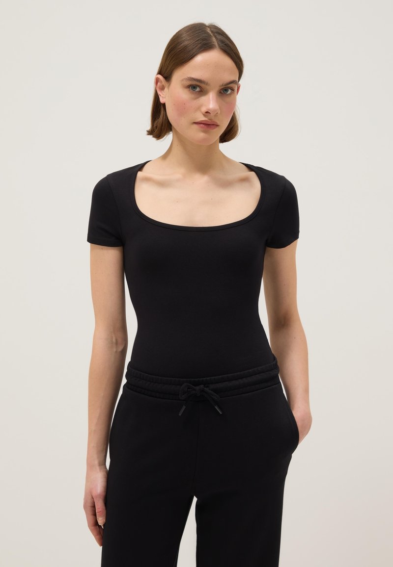 Jonge vrouw met kort bruin haar, gekleed in een aansluitend zwart T-shirt met korte mouwen en een ronde hals, en zwarte broek met trekkoord, staand tegen een effen achtergrond.