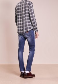 Kostkovaná košile s dlouhým rukávem v tmavě šedé a bílé barvě, spárovaná s modrými džíny a burgundskými botami. Pozorováno zezadu, ukazující volný střih.