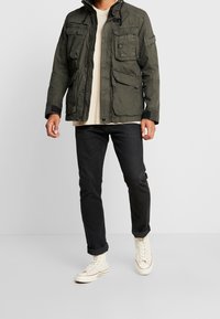 Olivgrön jacka med flera fickor, dragkedja och knäpplås, samt hög krage. Bärs över en beige skjorta och svarta jeans med vita sneakers.