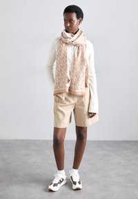 Beige scarf med repetitivt "KM" mönster, krämfärgad ribbad långärmad topp, beige shorts och slitna vita sneakers med mörka accenter.