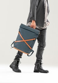 Zaino blu a rotolo con cinghie arancioni, realizzato in materiale resistente all'acqua. Presenta un logo e due modalità di trasporto. Design durevole.