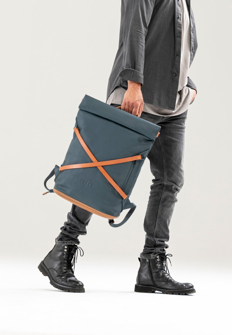Zaino blu a rotolo con cinghie arancioni, realizzato in materiale resistente all'acqua. Presenta un logo e due modalità di trasporto. Design durevole.