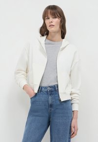 Haut zippé crème avec capuche, poches avant et poignets côtelés, superposé sur un t-shirt gris. Associé à un jean bleu taille haute.