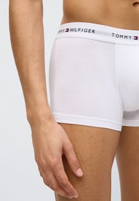 Λευκά βαμβακερά μποξέρ με αντίθετη μπλε σκούρα και κόκκινη ζώνη μέσης με την επιγραφή "TOMMY HILFIGER". Κοντινή λήψη των μποξέρ και χεριού.