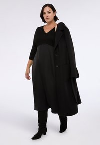 Černé šaty s výstřihem do V, zdobené cvočky v pase, doplněné dlouhým černým kabátem. Modelka má na sobě černé vysoké boty.