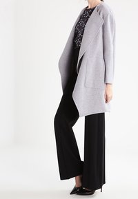 Vrouw in een lichtgrijze open jas over een bloempatronen top, zwarte brede broek en zwarte puntige hoge hakken tegen een witte achtergrond.