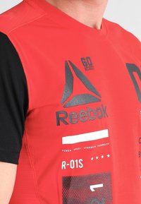 Camiseta deportiva de manga corta en rojo y negro, que presenta el logo de Reebok impreso, varios elementos de texto y un patrón de malla en la manga negra.