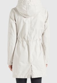 Giacca beige chiaro con cappuccio, vita cinchata e dettagli elastici. Presenta un tessuto liscio e opaco e un logo vicino al collo.