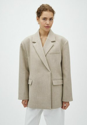 Jeune femme aux cheveux bouclés porte un blazer beige oversize et un pantalon blanc, debout devant un fond clair uni.
