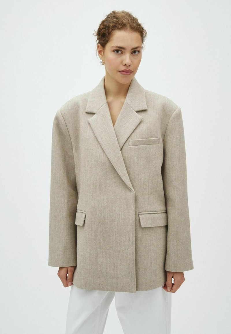 Jeune femme aux cheveux bouclés porte un blazer beige oversize et un pantalon blanc, debout devant un fond clair uni.