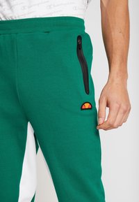 Gröna mjukisbyxor med svart dragkedjeficka på låret och orange-röd logotypapplikation, bärs av en person i vit Champion-tröja.