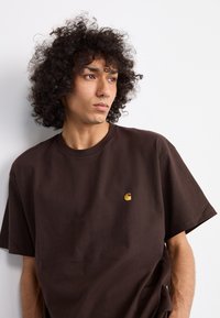 T-shirt in cotone marrone con vestibilità rilassata, dotato di un piccolo logo dorato sul lato sinistro del petto. Il tessuto appare liscio con una consistenza morbida.