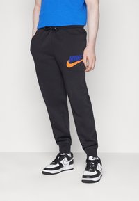 Svarta sweatshirtbyxor med en orange och blå Nike-logga, dragsko i midjan och ribbade muddar, tillsammans med svarta och vita Nike-sneakers.