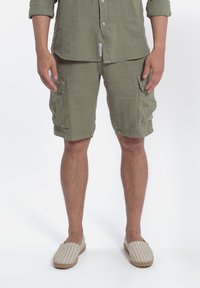 Camicia in lino e pantaloni cargo color verde oliva, con tasche applicate. Espadrillas beige a righe con suola intrecciata. Vestibilità pulita e casual.