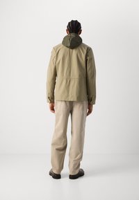 Jachetă khaki cu capușă verde, având talie ajustată, buzunare cu fermoar și mâneci lungi. Asortată cu pantaloni de un bej deschis și pantofi negri.