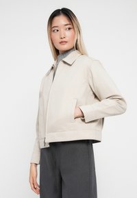 Béžová skrátená bunda s špicatým golierom, predným zipsom a bočnými vreckami. Tkanina má hladkú textúru; nosená cez sivý top.