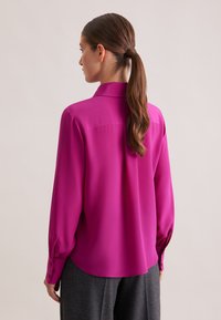 Blusa di seta fucsia con maniche lunghe, colletto a punta e polsini con bottoni. Texture liscia con vestibilità ampia e orlo curvo.