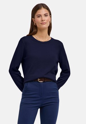 Femme aux longs cheveux bruns portant un pull bleu marine, un pantalon assorti, une ceinture marron, des boucles d'oreilles en forme d'anneau doré et un collier en or, debout avec les mains derrière.