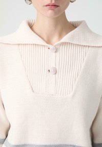 Dzianinowy sweter w jasnoróżowym kolorze z prążkowanym kołnierzem, z dwoma ozdobnymi guzikami i teksturowanym zaokrągleniem. Subtelne szare paski na dole.