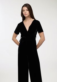 Czarny aksamitny kombinezon z krótkimi rękawami, głębokim dekoltem w serek, pasa z talią oraz szerokimi nogawkami, charakteryzujący się gładką, miękką teksturą i eleganckim designem.