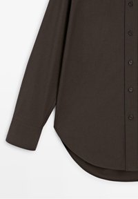Chemise marron foncé à manches longues avec poignets boutonnés, boutons sur le devant et ourlet arrondi, présentée sur fond blanc.