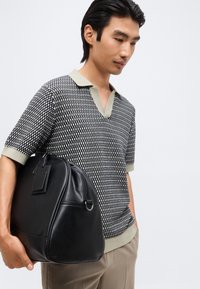 Νέος άνδρας με μπλούζα πόλο με σύντομο μανίκι με σχέδια και μπεζ παντελόνι κρατώντας μια μαύρη δερμάτινη τσάντα σε λευκό φόντο.