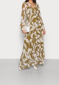 Maxi mekko oliivinvihreänä, jossa on valkoisia kukkakuoseja, pitkät hihat ja röyhelöhelma. Yhdistettynä pieneen valkoiseen käsilaukkuun ja vaaleansävyisiin kenkiin.