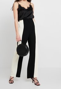 Kvinna som bär en svart cami med spetskant, högmidjade svartvita vida byxor, svarta klackar med remmar, och håller en rund svart handväska.