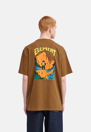 Persoon met kort rood haar die een bruine T-shirt draagt met een kleurrijke wandelcartoon en de tekst "ELEMENT" op de achterkant.