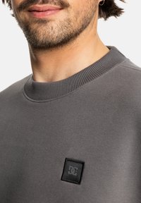 Sudadera gris de cuello redondo hecha de tejido suave, con un parche de logo negro cuadrado en el lado izquierdo del pecho y puños y cuello acanalados.