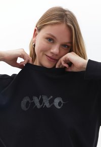 Czarna bluza z długim rękawem, okrągłym dekoltem i miękkim materiałem, ozdobiona błyszczącym srebrnym logo "OXXO". Minimalistyczny design.