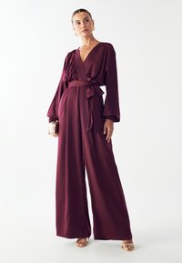 Bordo kombinezon z globokim V-izrezom, dolgimi rokavi in vezanim pasom. Narejen iz gladkega materiala z širokimi nogavicami in subtilnim sijajem.