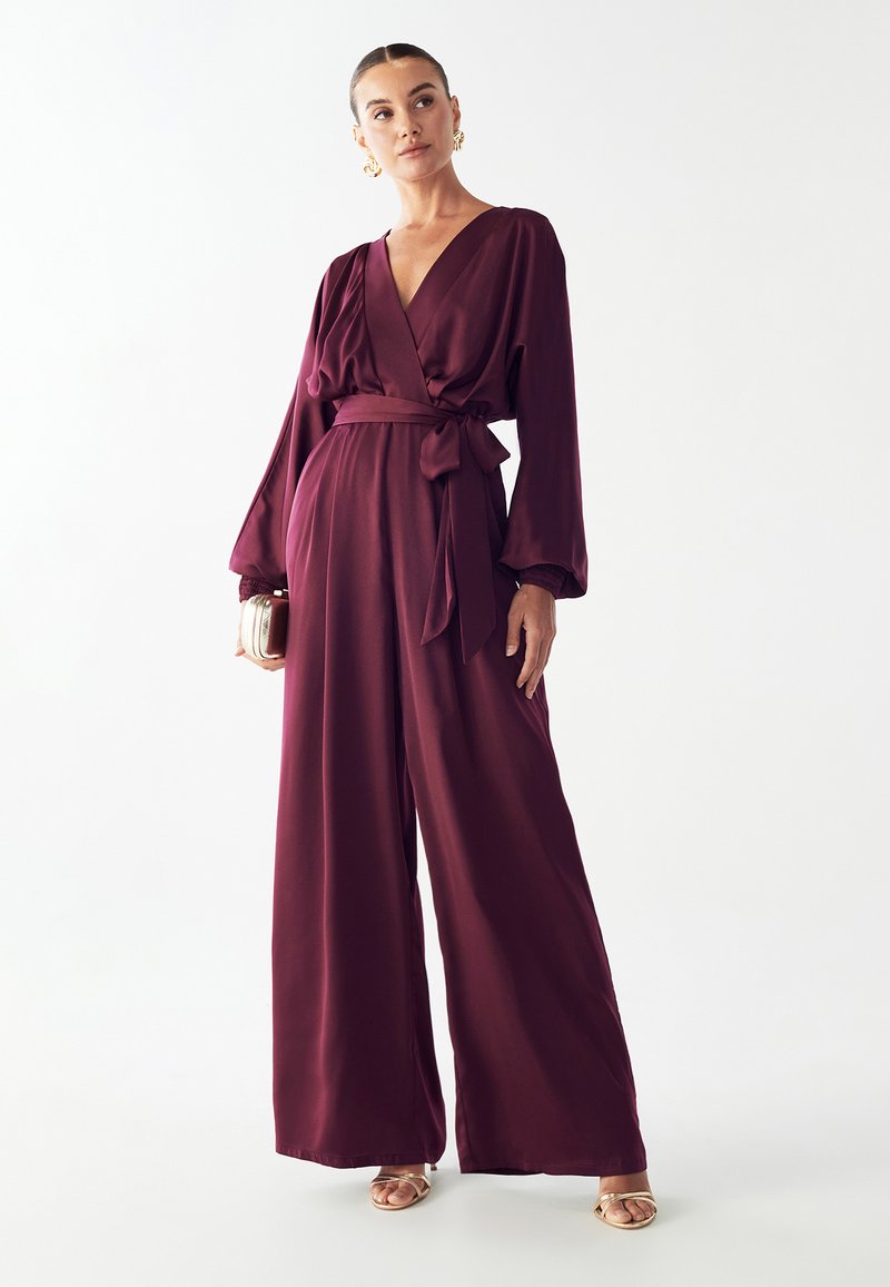 Bordo kombinezon z globokim V-izrezom, dolgimi rokavi in vezanim pasom. Narejen iz gladkega materiala z širokimi nogavicami in subtilnim sijajem.