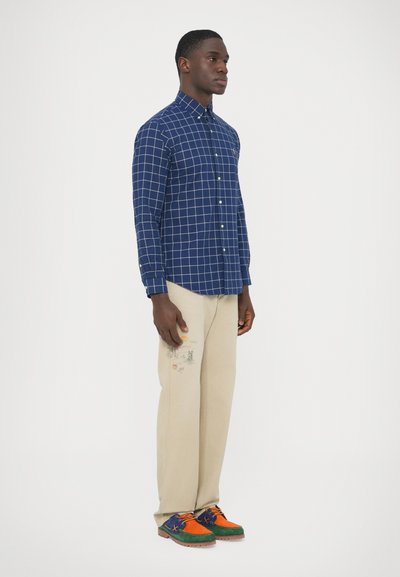 Mornarsko modra srajca s karirastim vzorcem in gumbi, bež hlače z živahnimi vezeninami ter večbarvni semišasti čevlji z oranžnimi, zelenimi in modrimi poudarki.