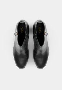Stivali alla caviglia in pelle nera con una texture liscia, punta affusolata e cerniera laterale, caratterizzati da dettagli in oro e design minimalista.