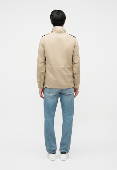 Homme de dos portant une veste beige, un jean bleu clair et des baskets blanches sur un fond blanc uni.