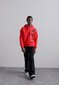 Sweat-shirt à capuche rouge avec le mot "ICON" sur le devant, associé à un pantalon noir et des chaussures à semelles blanches. Tissu lisse, coupe décontractée, design simple.