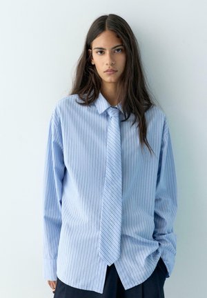 Mlada ženska z dolgimi temnimi lasmi, oblečena v veliko svetlo modro srajco s črtami in pripadajočo kravato, stoji pred preprostim belim ozadjem.