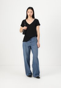 Black short-sleeve V-neck top paired with blue wide-leg jeans. Model holds a small black bag; outfit features a relaxed fit and lightweight fabric.