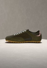 Χαμηλού προφίλ παπούτσι σε ελιά πράσινο με πλέγμα στα πλαινά, λευκά κορδόνια, ροζ λεπτομέρεια στη φτέρνα και υφή στο ανοιχτό καφέ πέλμα, πάνω σε ανοιχτή επιφάνεια.