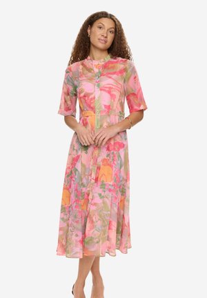 Kvinde iført en farverig blomsterprintet midikjole med knapper foran og ærmer til albuerne, stående med hænderne blidt foldet.