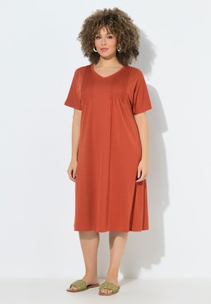 Kvinde med krøllet hår iført en rustfarvet knælængde kjole med pikkut detaljer og grønne vævede åbne tå-sandaler, stående mod hvid baggrund.