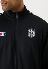 Jachetă sport neagră cu fermoar, cu guler înalt, având accente albe și un logo al unui club de fotbal pe pieptul stâng.
