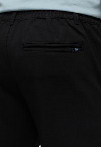 Nærbilde av baklomme på svarte bukser med en liten svart og hvit stofflogo på høyre side, brukt med en lyseblå skjorte.