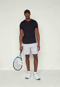 Μαύρο αθλητικό μπλουζάκι, γκρίζες βερμούδες, κρατώντας μια ρακέτα τένις. Φοράει λευκά αθλητικά παπούτσια και έναν αντίστοιχο κορδέλα. Απλός, λειτουργικός σχεδιασμός.