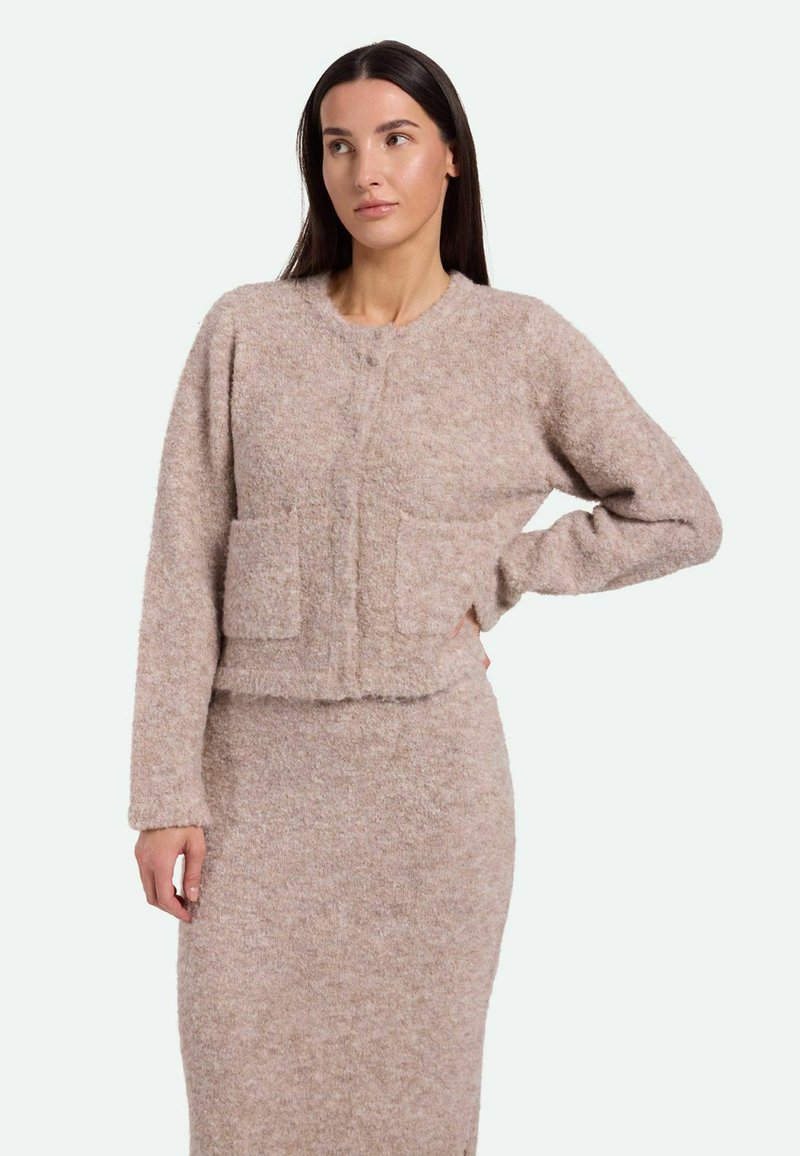 Gekürzter, fusseliger beiger Cardigan mit rundem Ausschnitt, Knopfverschluss vorne und zwei aufgesetzten Taschen. Weicher Stoff und minimalistisches Design.
