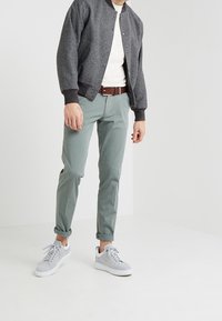 Giacca bomber grigia, maglietta a girocollo bianca, pantaloni chino verdi, cintura marrone e sneakers grigie chiare con accenti bianchi.